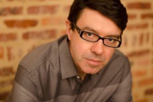 man wit brown hair black glasses gray shirt by brick wall