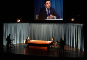 2 people seated at long table by curtains with tv screen overhead