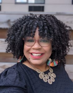 Soraya Nadia McDonald smiling with black curly hair, red glasses and colorful jewelry