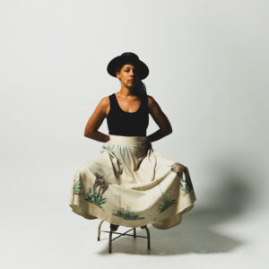 kamara thomas wears black hat, tank and white skirt while seated on stool