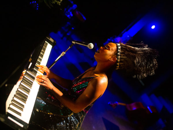 tanyaradzwa plays piano and sings at mic, wearing sequined dress and dark feathered headdress