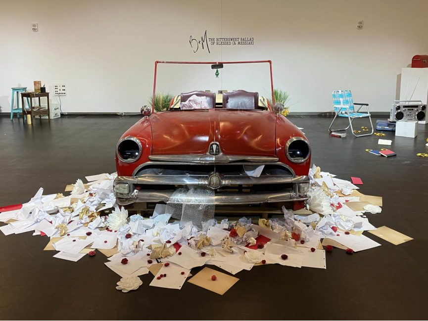 A vintage car surrounded by paper sits in an art gallery