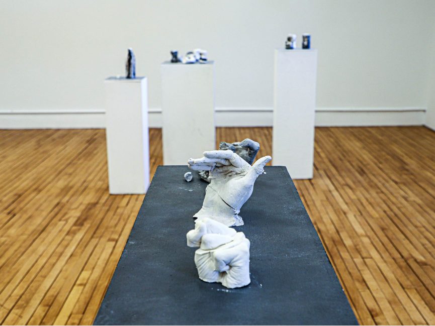 sculptural work of white gestural hand rests on dark table, other works seen on podiums behind