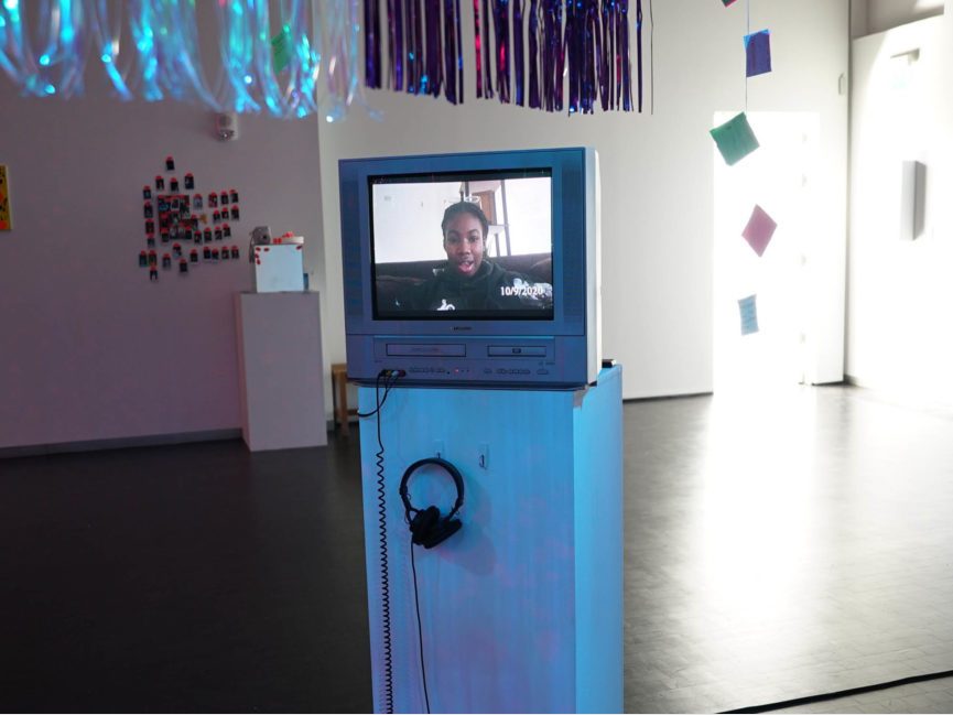 A TV sits on a pedestal with an image of a person is on the screen. Headphones on the pedestal hang from a hook