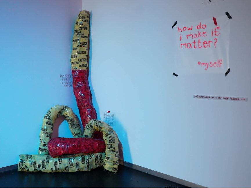 A tube like sculpture covered in caution tape is displayed in the corner