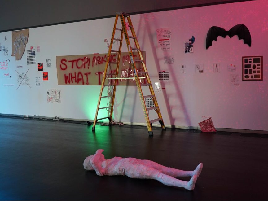 A figure lies on the floor with as a ladder stands tall in the background against a wall. The wall is covered by various art