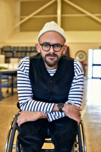 ryan gander sits in wheelchair, wears white beanie, striped shirt and black glasses