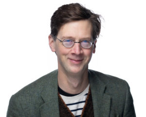 manchan magan smiles at the camera. He has reddish brown short hair, glasses, and wears green blazer with red vest and striped shirt.