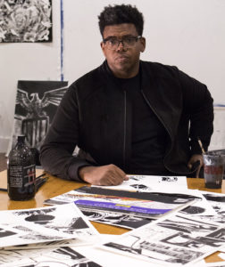 mark thomas gibson wears black jacket and gazes directly at camera with one hand on table, one on his chair arm. black and white ink drawings scattered on tabletop in front of him.