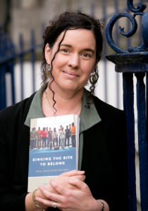 helen pheland stands near wrought iron gate holding a book. she has dark brown curly hair and wears a green collar shirt and black blazer