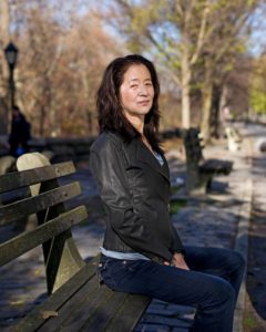 julie otsuka sits on a bench in a park. She sits in profile and looks at camera, wearing leather jacket and jeans