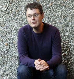 Ilya Kaminsky sits against a rock wall wearing a purple sweater and rimless glasses, looking slightly off to the side.