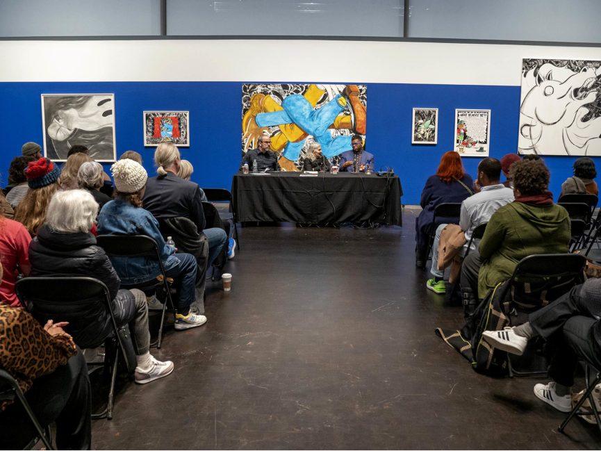 Three artists sit at a table discussing art with an audience