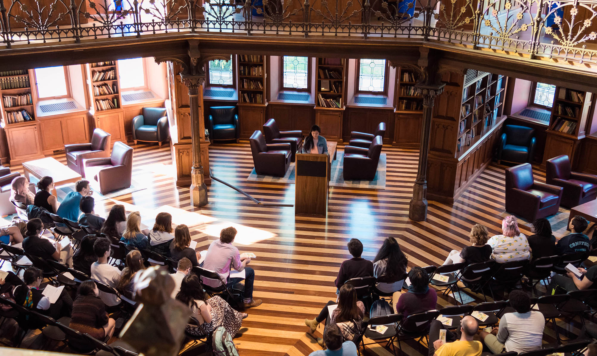 one student reads at a podium in front of a seated crowd in a grand library with striped wooden floors and ornate trim work