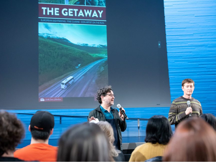 A poster for "The Getaway is projected behind two people talking to an audience