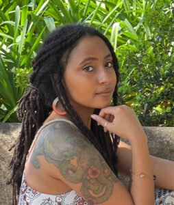 Dantiel Moniz sits in profile and looks over her right shoulder with chin resting on fingers. She has long dark braided hair and a large colored tatoo on her left shoulder.