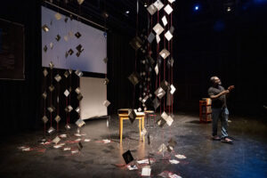 Dwayne Betts stands onstage by a small table surrounded by hanging red threads filled with paper kites designed by Kyoko Ibe.