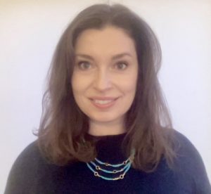 geraldine parsons smiles with straight brown hair to her shoulders. she wears a blue beaded three strand necklace and navy sweater