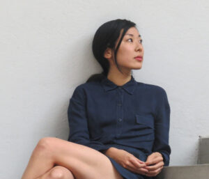 Katie Kitamura sits with legs crossed, reclined against white wall. She wears a navy collar button up shirt and short skirt. She has dark hair pulled back and looks off to the right.