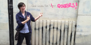 Lyra McKee stands and smiles and points by a stone wall written with grafitti.