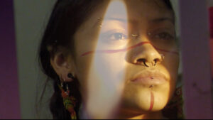 A woman stares off in the distance, seen closeup with red paint lines and piercings on her face.