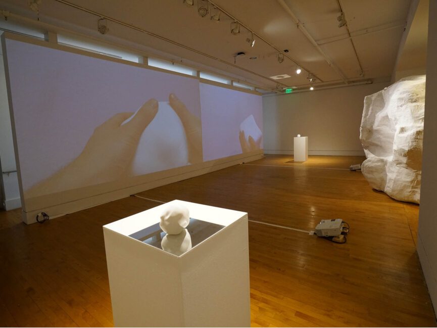 A small cube like sculpture sits on a pedestal in a gallery with a projection of hands manipulating the cube in the background