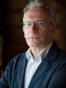 Fintan O'Toole gazes seriously at camera. He has short gray hair, wears rectangle wire glasses, and wears a white collared shirt and navy blazer.