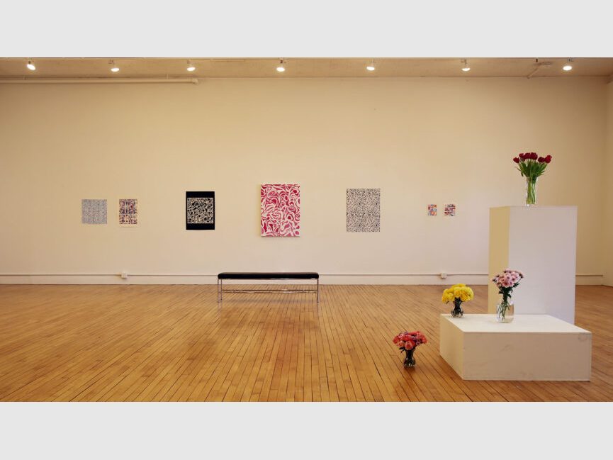 sunlit gallery space with a black bench, colorful works on walls, and flowers on podiums