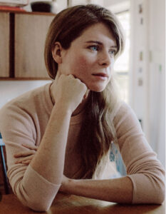 Emma Cline looks off to left with head resting against her right knuckles. She sits in a sunlit room.