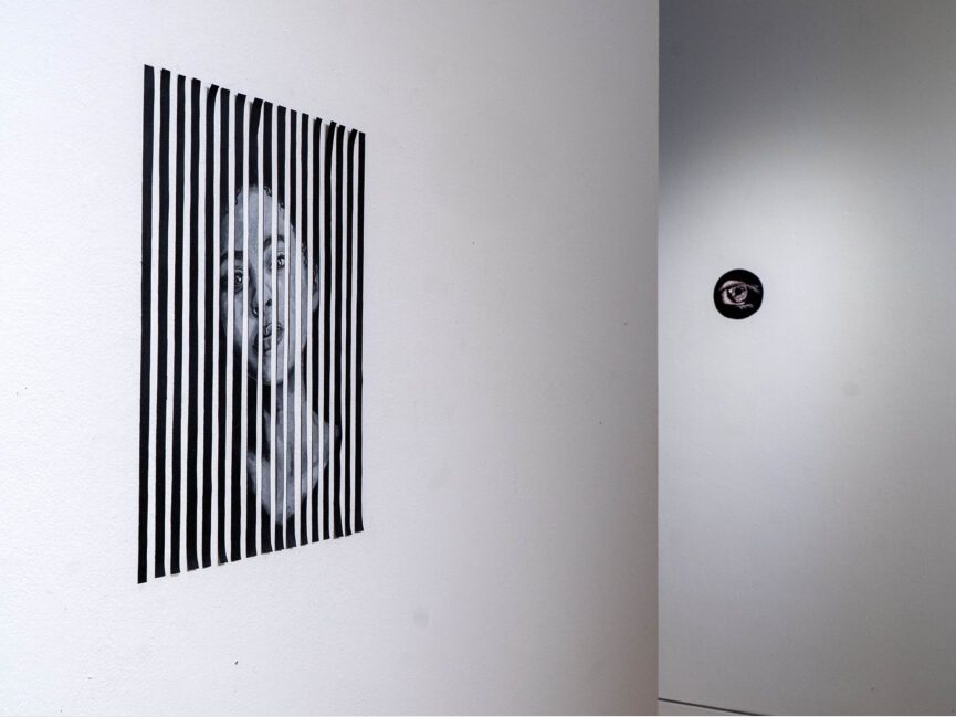 A small painting of an eye peeks around a corner as a larger painting of a person staring is front and center.