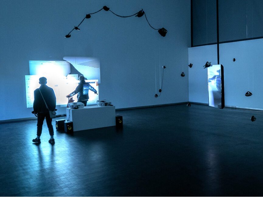 Two people walk through a gallery lit in a blue light screens and objects hanging from the ceiling.