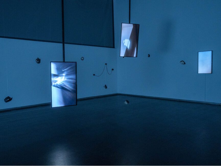 Screens and objects hang from the ceiling in a very dark gallery.