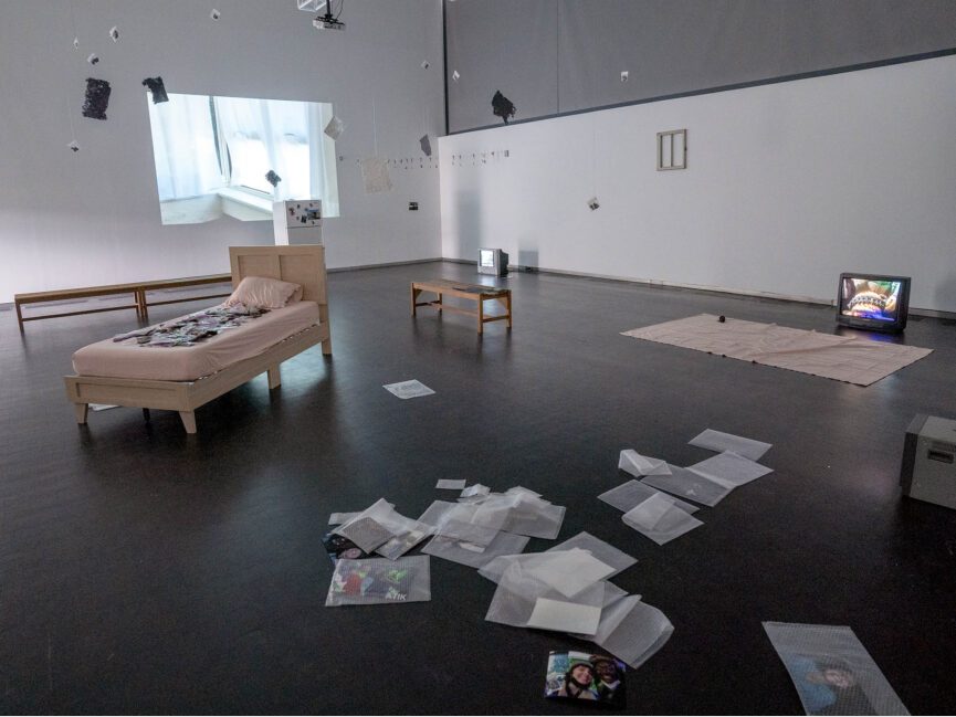 elements of apartment furniture and phtoos scattered in large open gallery space, with video projection on wall