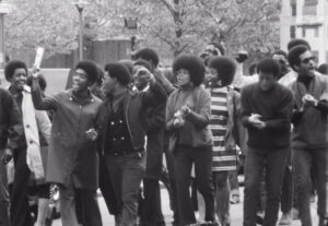 Historical black and white photo of a group of Black students walks together outside, some clapping hands and some raising fists triumphantly
