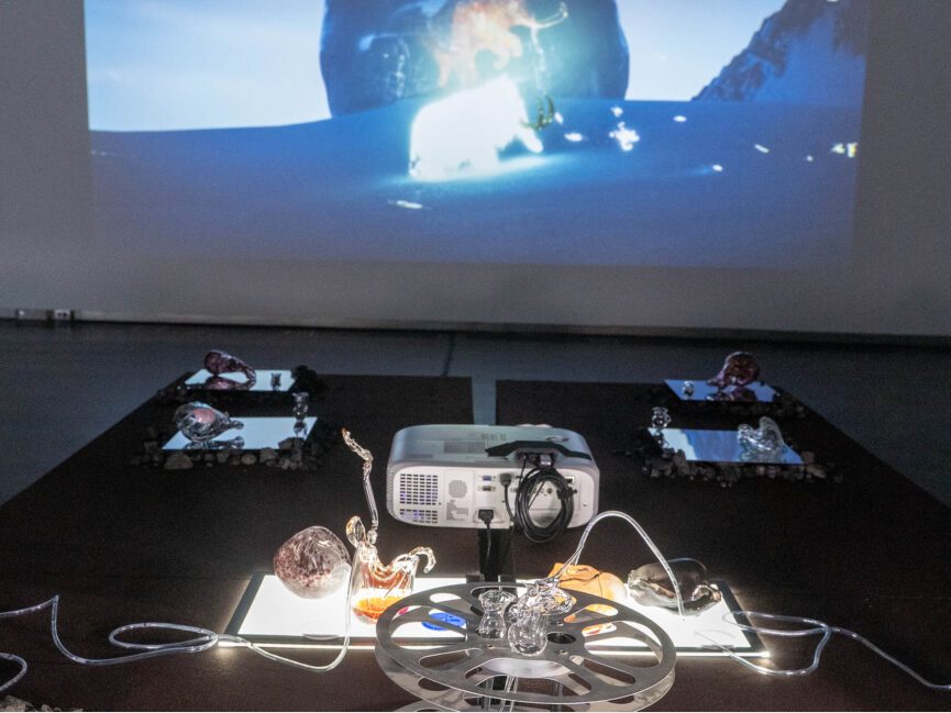 Translucent artifacts are on view on a table as a projection of a mountain scape is seen in the background