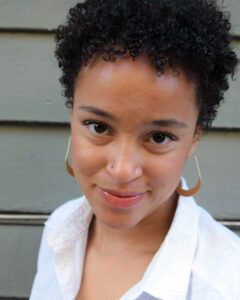 Tanaka offers a closed-lip smile. She wears large hoop earrings and has short dark curly hair.