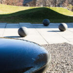 A round, low fountain filled with water in the foreground seen with spherical sculptures, sidewalk and grassy hills in background.