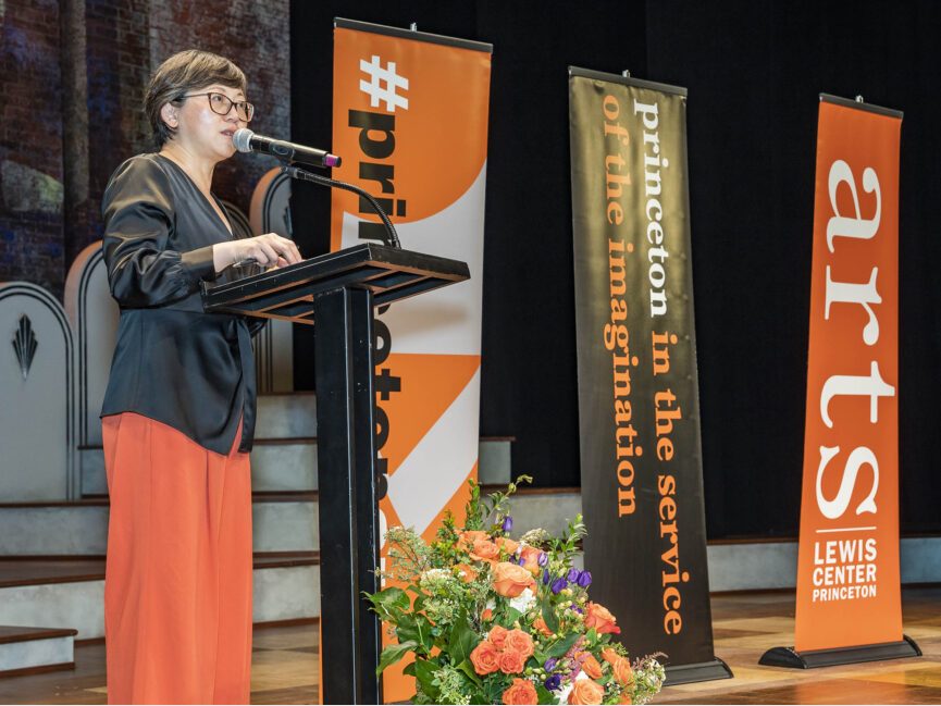 A person talks into a microphone at a podium as orange a black banners promoting the arts program stand beside her