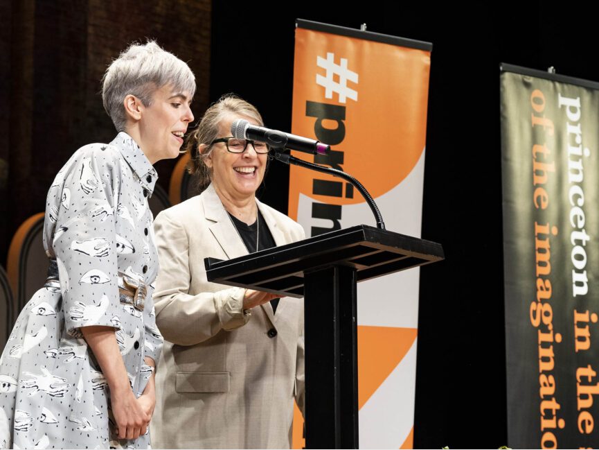 Two people speak at a mic behind a podium besides orange and black banners