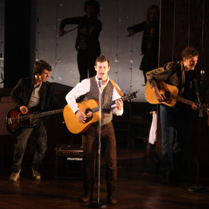 Sam Gravitte stands playing guitar and singing at a mic onstage with other musicians behind him.