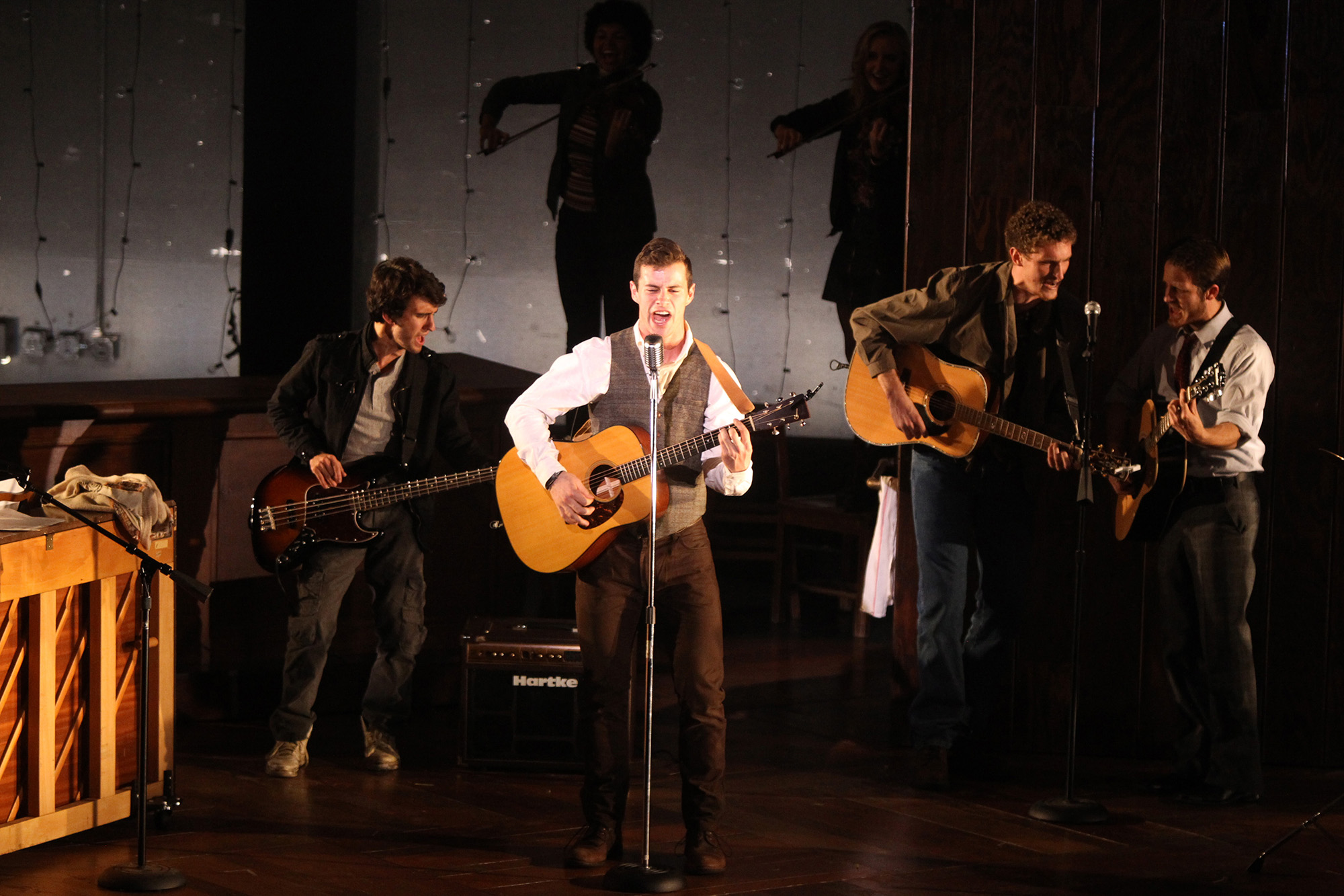 Sam Gravitte stands playing guitar and singing at a mic onstage with other musicians behind him.