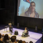 Two people onstage talk with a person on a Zoom screen projected behind them. ASL Interpreters present on the left side of the stage.