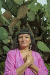 Writer Sandra Cisneros stands outside by a tall green and brown cactus.