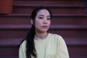 Writer Ada Zhang looks at the camera. She wears a black necklace at her throat and a long sleeved yellow tshirt.