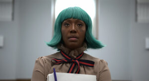 A black person with an aqua green wig faces the camera. They wear a red and navy bow tied at the neck of a high collared brown shirt.