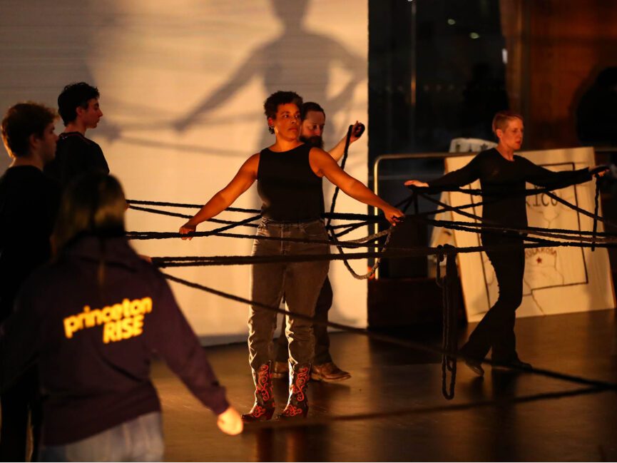 Kamara Thomas and other performers hold and stretch out several black ropes.