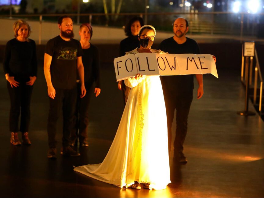 An actor holds a sign that reads follow me in front of a group of other performers.