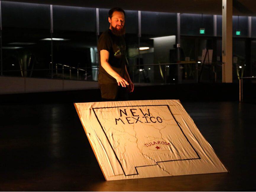 A performer on stage points to a crude map of New Mexico with Tularosa highlighted.