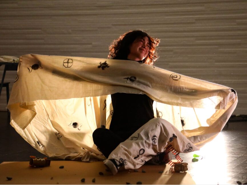 A performer wrapped in a sheet covered in symbols appears to be rising from a deep sleep.