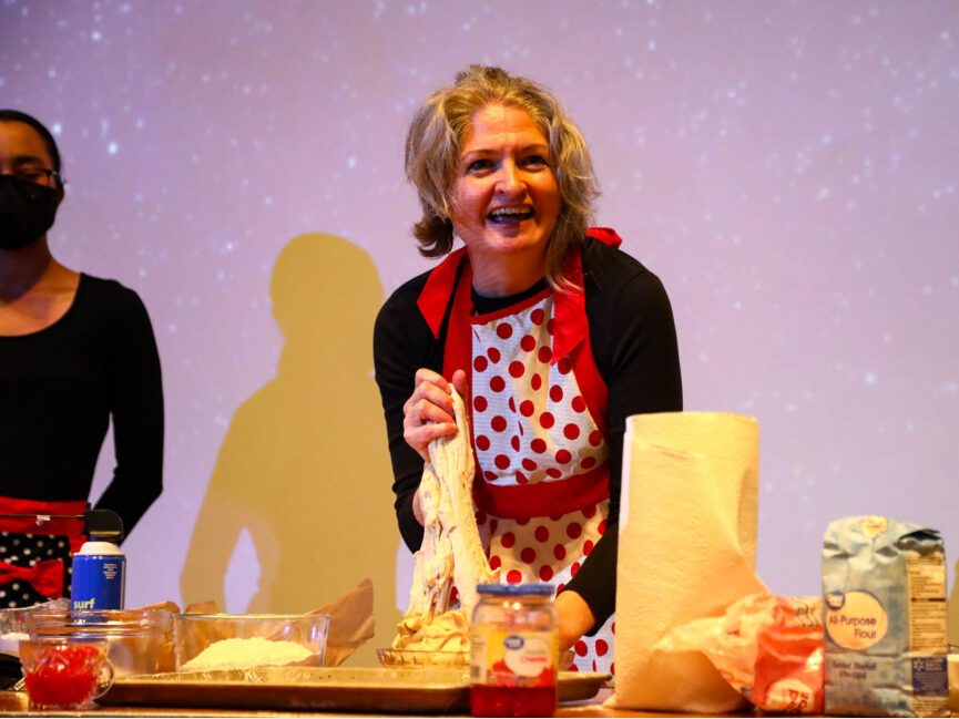 A performer wearing a colorful apron makes something at a cluttered table